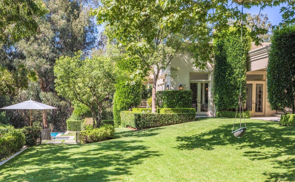 A lawn and trees in the foreground and a house with glass doors and a nearby pool are in the background. 